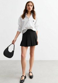 Chemise blanche à rayures avec une coupe décontractée, associée à une jupe noire plissée. Sac à main noir à forme courbée, et chaussures plates noires avec des sangles.