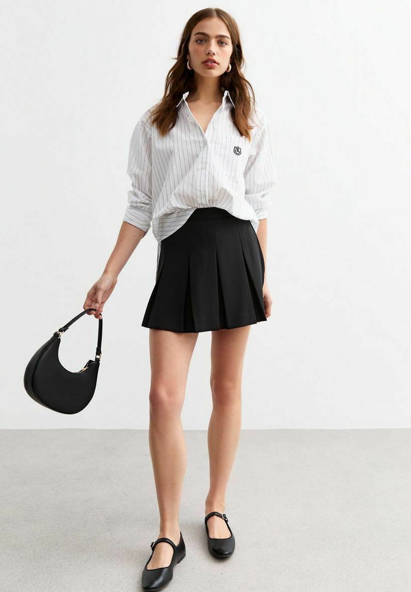 Chemise blanche à rayures avec une coupe décontractée, associée à une jupe noire plissée. Sac à main noir à forme courbée, et chaussures plates noires avec des sangles.