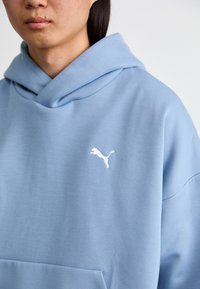 Lysblå hættetrøje lavet af blødt stof med en stor frontlomme. Har et hvidt Puma-logo på venstre brystområde og en snoretrækshætte.