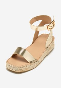 Sandalia de espadrille de cuero dorado con tiras anchas, tira de tobillo y detalle de hebilla. La suela de yute trenzado añade textura y contraste.