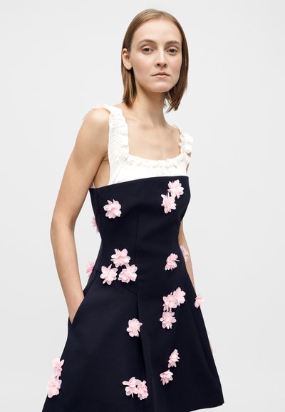 Abito blu navy con scollo quadrato e spalline bianche arricciate, decorato con fiori di stoffa rosa sulla gonna e tasche laterali. Tessuto dalla trama liscia.