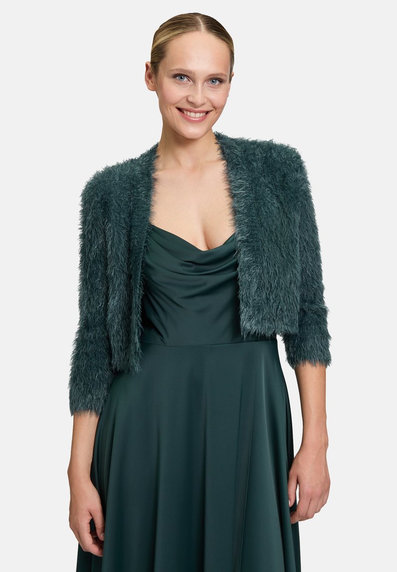 Faux-Fur, dunkel-türkisfarbene, kurze Jacke mit langen Ärmeln, die eine weiche Textur und ein offenes Frontdesign aufweist, getragen über einem passenden Kleid.