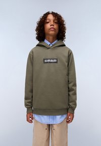Olivgrüner Hoodie mit einem Logo-Patch auf der Vorderseite, getragen über einem blau-weiß gestreiften Hemd. Der Stoff wirkt weich und strukturiert.