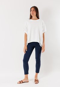 T-shirt bianca oversize a maniche corte, abbinata a pantaloni slim-fit blu navy. Il modello indossa sandali marroni. Lo sfondo è completamente bianco.