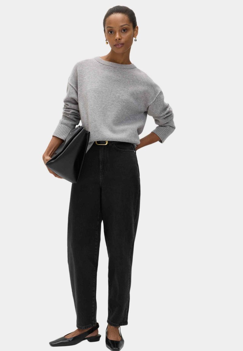 Suéter de punto gris con mangas anchas, cuello alto y puños acanalados, combinado con jeans negros de tiro alto y zapatos planos negros.