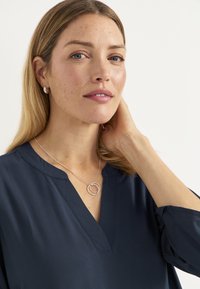 Silverhalsband med sammanlänkade cirkulära hängen, bärs över en marinblå blus med V-ringning och mjuk tygtextur.