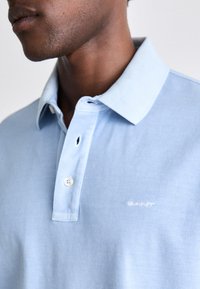 Man draagt een lichtblauw poloshirt met twee vastgemaakte knopen, met kraag en vaag wit logo op de borst.