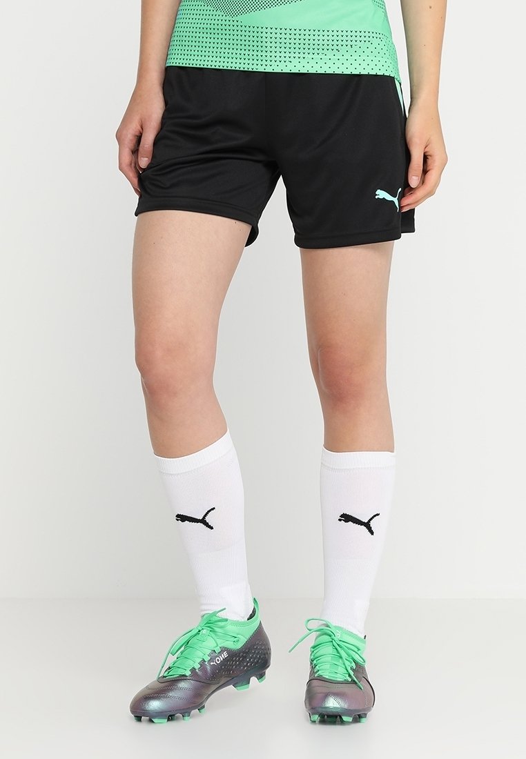Personne portant un short noir Puma, des chaussettes blanches Puma jusqu'aux genoux et des chaussures de football noires et vertes, debout sur un fond uni.