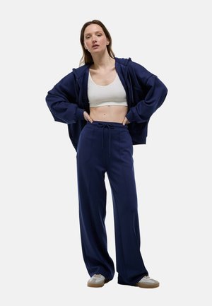 Sweat à capuche oversized bleu marine et pantalon large assorti en tissu doux, associés à un crop top ajusté blanc et des baskets légères.