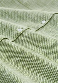 Tessuto verde texturizzato con motivo a griglia, caratterizzato da bottoni bianchi. Il materiale sembra leggero e leggermente stropicciato.