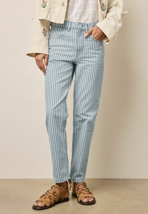 Frau trägt hoch taillierte blau-weiß gestreifte Hosen, beigen bestickten Blazer und braune Schnürsandalen, steht mit einer Hand in der Tasche.