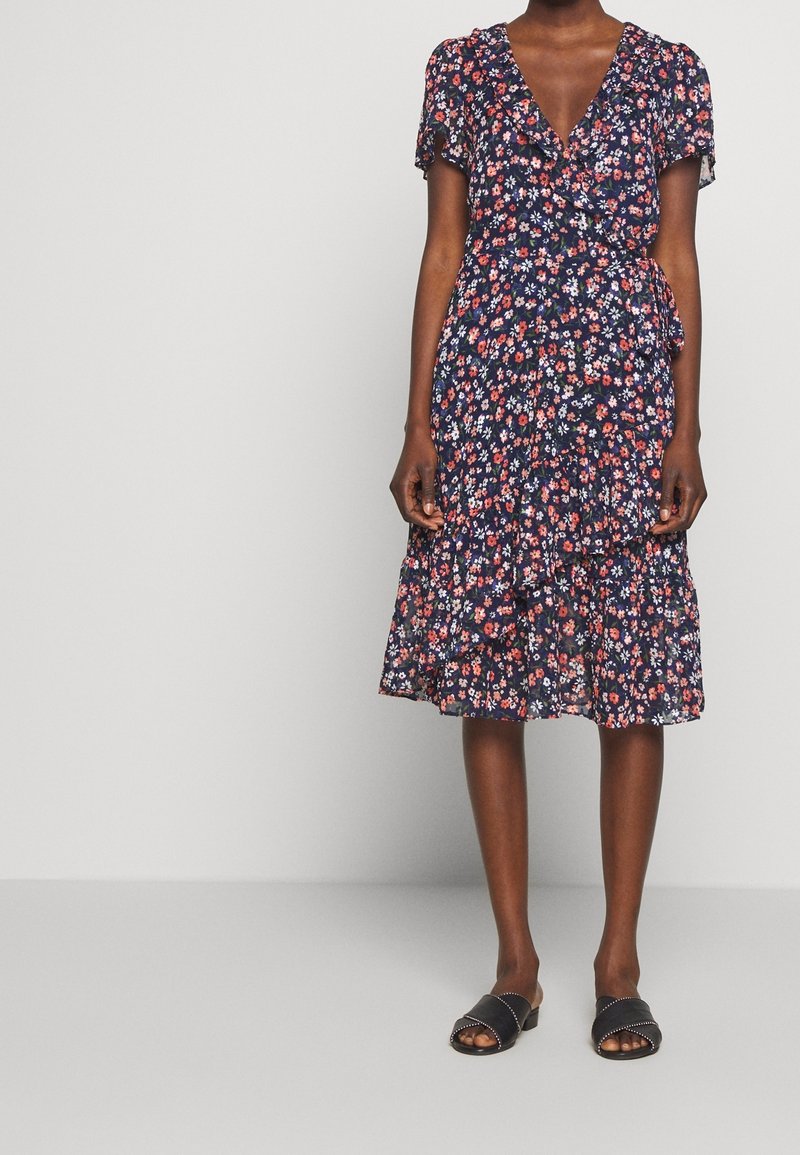 Femme portant une robe portefeuille à fleurs bleu marine à manches courtes et des sandales noires ouvertes à enfiler, debout devant un fond gris clair.