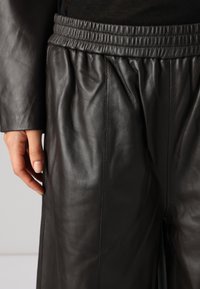 Zwarte leren broeken met een relaxte pasvorm, voorzien van een elastische tailleband en subtiele verticale plooien voor textuur en beweging.