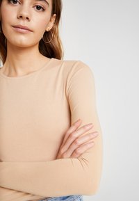 Beež pikka varrukatoppi pehme tekstuuriga, ümmargune kaelus ja keha järgiv disain. Modelil on käed ristis, näidates kangast üksikasju.
