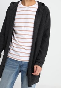 Kardigan in Kohlegrau mit Kapuze und langen Ärmeln, aus strukturiertem Stoff; getragen über einem weißen Shirt mit hellrosa horizontalen Streifen.