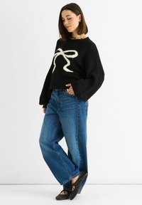 Maglione corto nero con design a nastro bianco, abbinato a jeans blu a vita alta e ballerine nere. Materiale in maglia morbido, vestibilità comoda.