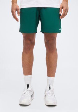 Homme portant un short de sport vert, des chaussettes blanches et des chaussures de course blanches, debout sur un fond uni.