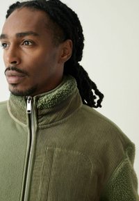 Olivengrøn jakke med lynlåslukning ved kraven, en kombination af bløde og ribbede teksturer, med en brystlomme og sølvfarvet lynlåsdetalje.