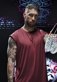 Maglietta sportiva rossa senza maniche con un logo nero, tenuta da una persona tatuata che indossa scarpe da calcio bianche e dorate con disegni intricati.