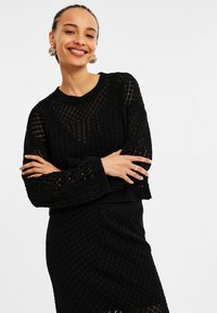 Schwarzer gestrickter Pullover mit offener Maschenstruktur, runder Halslinie und gerippten Bündchen, kombiniert mit einem passenden strukturierten Rock.