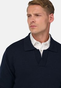 Jūras zila adīta virsjaka ar strukturētu audumu, kurai ir plata apkakle un pusplāksne virs baltas apkakles kreklā.