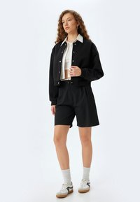 Giacca nera corta con bottoni argentati, camicia bianca con colletto e pantaloni corti neri. Calze lunghe fino alla caviglia con righe grigie e sneakers bianche con accenti neri.
