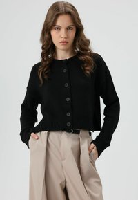 Zwarte cropped cardigan met een geribbelde zoom, voorzien van zes ronde knopen en lange mouwen, gecombineerd met crèmekleurige broeken. Gladde textuur.