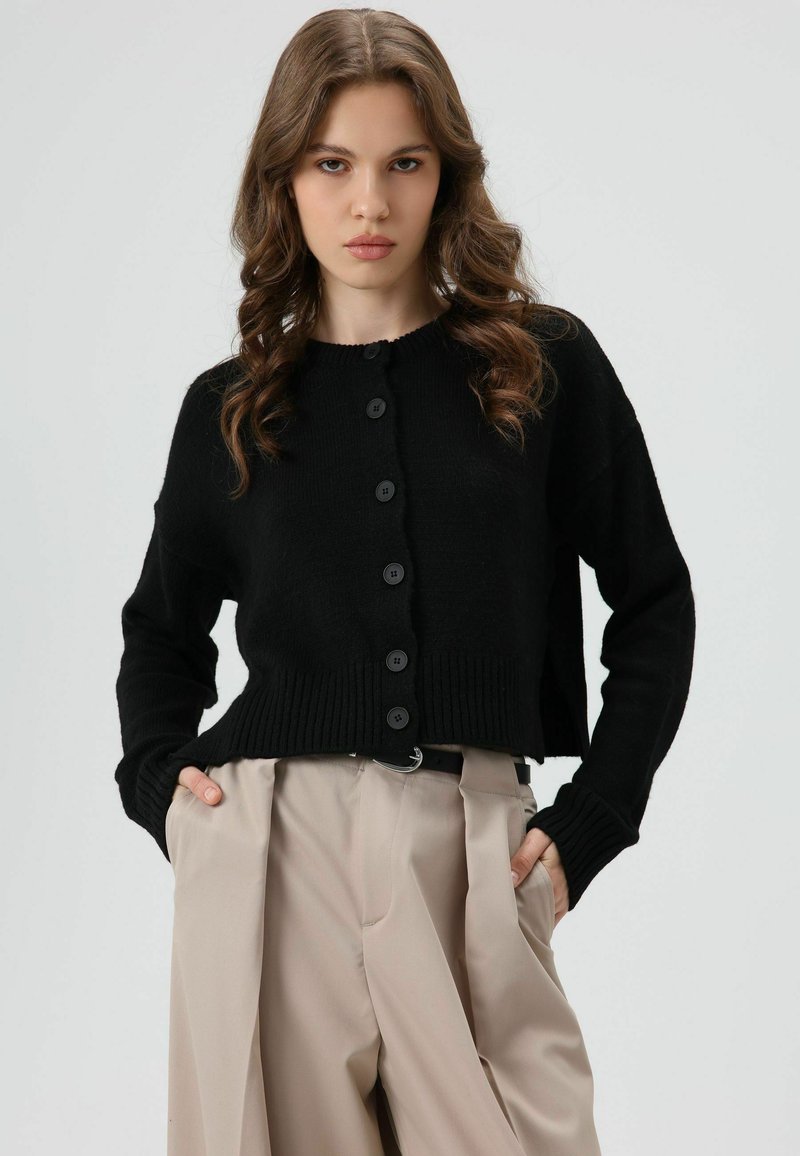 Zwarte cropped cardigan met een geribbelde zoom, voorzien van zes ronde knopen en lange mouwen, gecombineerd met crèmekleurige broeken. Gladde textuur.