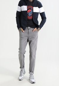 Giacca zip-up navy con strisce bianche, t-shirt grafica nera, jeans grigi e sneakers bianche, con maniche arrotolate e dettagli di tatuaggi.