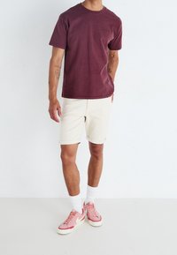 Muž v tmavě červeném tričku, krémových šortkách, bílých ponožkách a růžových teniskách Nike stojí proti jednobarevnému bílému pozadí.