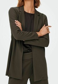 Blazer verde oliva con una vestibilità rilassata, maniche lunghe e apertura frontale. Abbinato a un top nero e pantaloni coordinati. Tessuto dalla texture liscia.