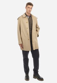 Beige esőkabát, gallérral, elöl gombokkal és két oldalsó zsebbel. Sötét polo póló és fekete nadrág felett viselve. Fekete fűzős csizma.