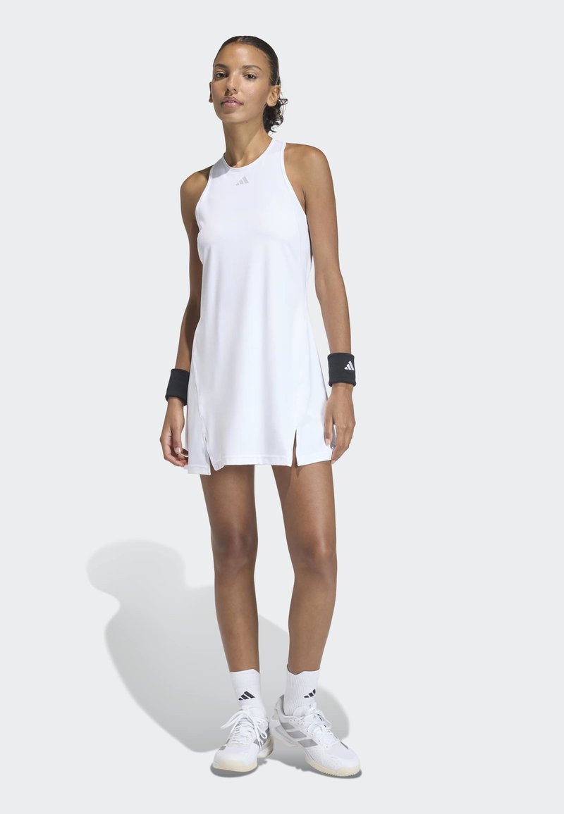 Mujer de pie vistiendo un vestido atlético blanco sin mangas, muñequeras negras, calcetas blancas y zapatillas blancas sobre un fondo claro y liso.