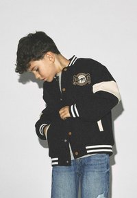 Veste de varsity noire avec des accents crème, des poignets rayés et un écusson de baseball à l'avant. Portée sur une chemise grise avec un jean bleu.