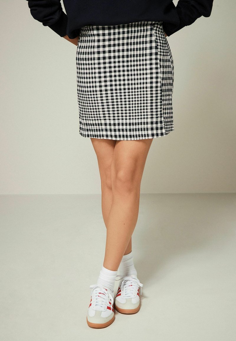 Jupe à carreaux noir et blanc avec une silhouette ajustée, associée à des baskets blanches rehaussées de rouge et des chaussettes blanches, se tenant sur un fond clair.