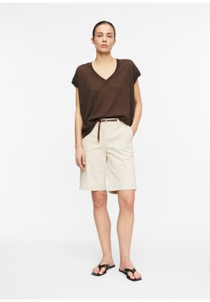 Kobieta stojąca, ubrana w luźny brązowy top z dekoltem w serek, beżowe spodenki do kolan z plecionym paskiem oraz czarne sandały na otwarte palce na białym tle.