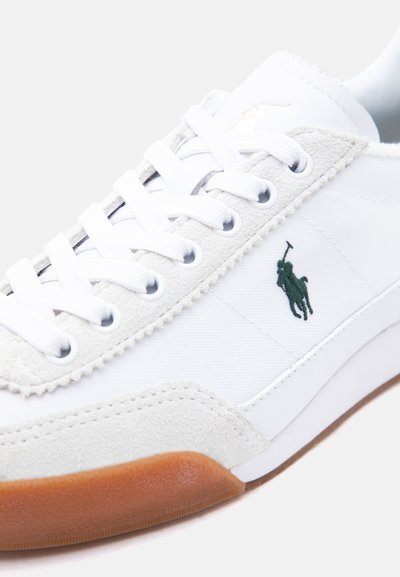 Fehér tornacipő velúr részletekkel, fehér fűzőkkel, gumitalppal és egy zöld hímzett póló játékos logóval az oldalon.