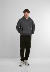 Grå huvtröja med känguruficka och lös passform, i kombination med svarta sweatpants. Modellen bär vita och blå sneakers.