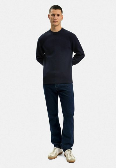 Marinblå stickad sweatshirt med rundad hals, kombinerad med mörkblå jeans och ljusa sneakers. Slät textur och enkel design.