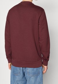 Sudadera color burdeos con mangas largas, puños acanalados y cuello redondo. Textura suave con un diseño sencillo, combinada con jeans de color azul claro.