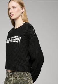 Schwarzer Strickpullover mit weißem "True Religion"-Schriftzug, Cropped-Design, geripptem Saum und Knopfakzenten an der Schulter. Kombiniert mit gemusterten grünen Hosen.