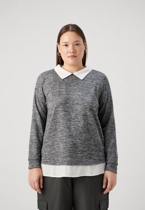 Jersey gris texturizado con cuello blanco, mangas largas y dobladillo redondeado, superpuesto a una camiseta blanca. Llevado con pantalones oscuros de estilo cargo.