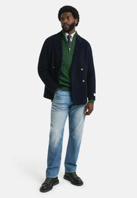 Marineblauer doppelt geknöpfter Mantel, grüner Grobstrickpullover, hellblaue Jeans, schwarze Schnürschuhe. Glatte Stoffe und lässige Passform.