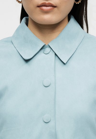 Γυναίκα που φορά χρυσούς κρίκους σκουλαρίκια και ανοιχτό μπλε σακάκι με γιακά και κουμπιά καλυμμένα με ύφασμα, κομμένο στο πηγούνι και τους ώμους.