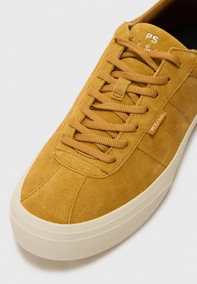 Mustárszín velúr sneaker lapos fűzőkkel, kerek orral és krémszínű gumitalppal. Oldalán egy kis márkacímke található.