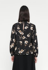 Svart bluse med hvit og beige blomstermønster, lange ermer, detalj med nøkkelhull bak, og innsamlede mansjetter, laget av et lett stoff.