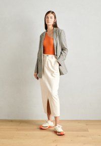Blazer in tessuto a quadri, canotta arancione, gonna crema con spacco frontale e sneakers beige con accenti arancioni. Texture morbide, vestibilità sartoriale.