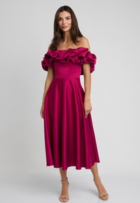 Sukienka w kolorze magenta z odkrytymi ramionami, z falbaniastym dekoltem, dopasowanym gorsetem i zwiewną spódnicą w kształcie litery A, uszyta z gładkiego materiału. Beżowe sandały na wysokim obcasie.
