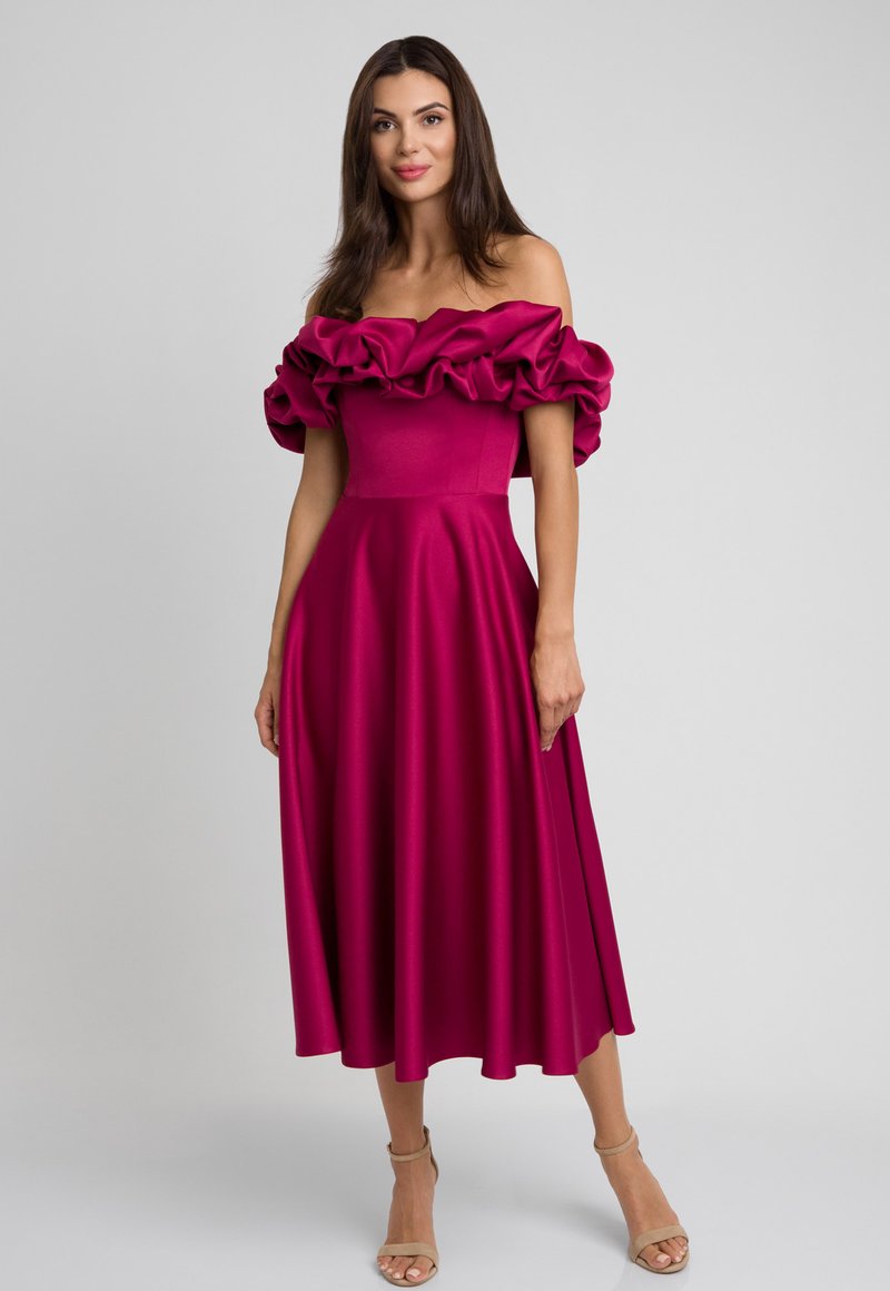 Sukienka w kolorze magenta z odkrytymi ramionami, z falbaniastym dekoltem, dopasowanym gorsetem i zwiewną spódnicą w kształcie litery A, uszyta z gładkiego materiału. Beżowe sandały na wysokim obcasie.