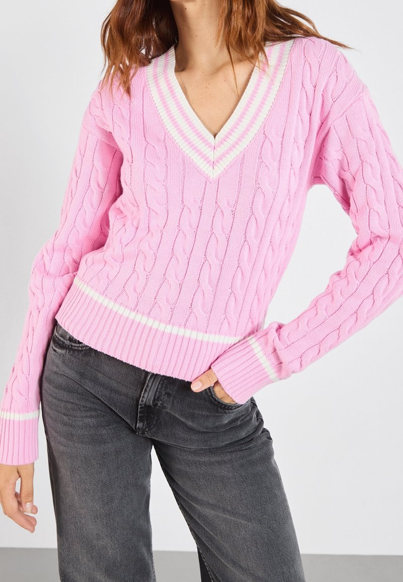 Femme portant un pull rose à col en V en maille torsadée avec des rayures blanches sur le col, les poignets et l'ourlet, associé à un jean gris foncé.